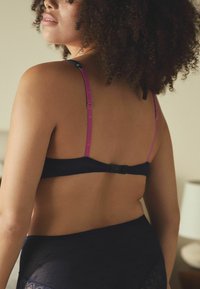 Sieviete ar kraukšķīgām matiem, kura valkā melnu krūšturi ar rozā siksnām un atbilstošu melnu mežģīņu apakšveļu, redzama no aizmugures maigi apgaismotā telpā.