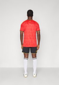 Κόκκινη αθλητική φανέλα με υφή φλόγας, μαύρα σορτς με κίτρινες λεπτομέρειες και λευκές κάλτσες. Περιλαμβάνει το λογότυπο της Adidas στα σορτς.