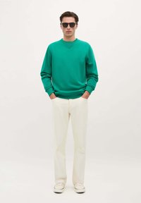 Groene sweatshirt met geribbelde manchetten, gecombineerd met crèmekleurige tapered broeken en lichtgekleurde sneakers. Model staat met de handen in de zakken.