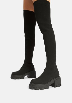 Bottes noires cuissardes avec talons blocs épais et semelles épaisses rainurées portées sur des jambes nues sur un fond uni.