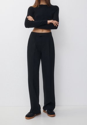 Femme debout, bras croisés, portant un top noir court à manches longues, un pantalon noir taille haute à jambes larges et des baskets noires avec des semelles marron.