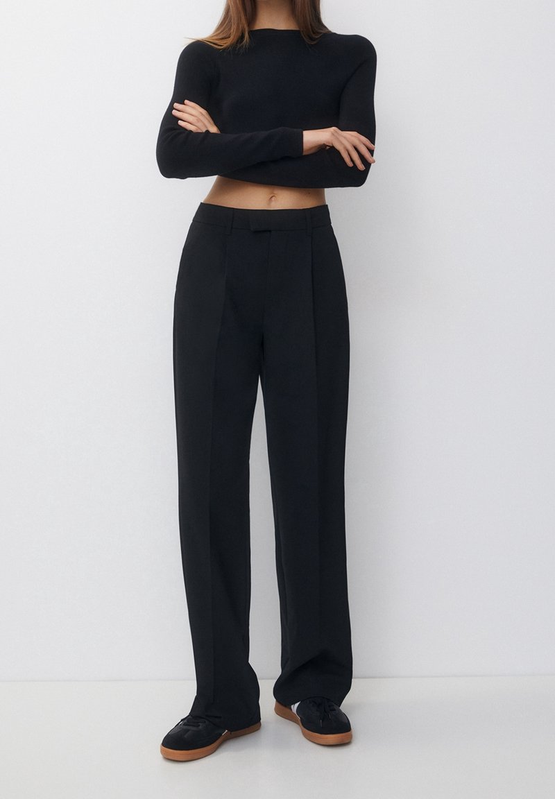 Femme debout, bras croisés, portant un top noir court à manches longues, un pantalon noir taille haute à jambes larges et des baskets noires avec des semelles marron.