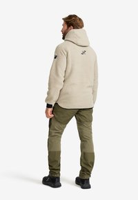 Beige fleecejacka med huva, prydd med en svart logotyp på baksidan. Kombinerad med olivgröna byxor och svarta skor.