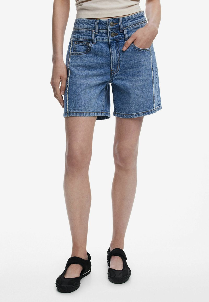Középkék farmer rövidnadrág, többutas magas derékkal, öt zsebbel és kontrasztvarrással. Fekete slip-on cipőkkel párosítva.