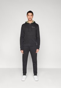 Μαύρο φούτερ με φερμουάρ, κορδόνια, μπροστινές τσέπες και λογότυπο στο στήθος, συνδυασμένο με μαύρο παντελόνι ίδιου χρώματος και λευκά παπούτσια.