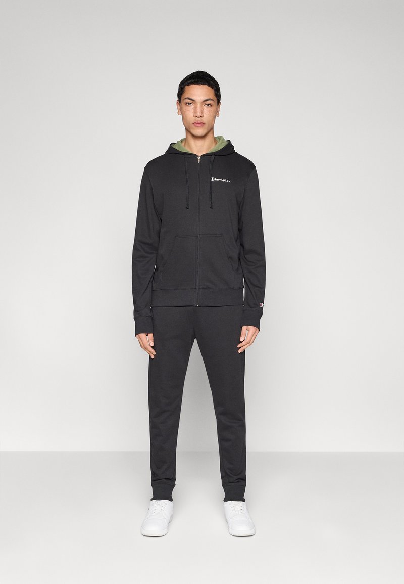 Μαύρο φούτερ με φερμουάρ, κορδόνια, μπροστινές τσέπες και λογότυπο στο στήθος, συνδυασμένο με μαύρο παντελόνι ίδιου χρώματος και λευκά παπούτσια.