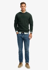 Zielona bluza z wypukłym logo „Superdry”, nałożona na niebiesko-białą koszulę w paski. Do tego jasne dżinsy i beżowe buty.