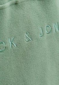Fleece tkanina v mint zelenkasti barvi s vezenim napisom "JOCK & JON" v svetlejši odtenku mint, z mehkobno in teksturirano površino.