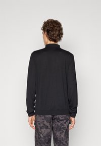 Camisa polo preta de manga longa com colarinho, tecido macio, punhos e barra canelados. Usada com calças escuras cinza padrão.