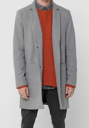 Man draagt een grijze wollen jas over een oranje gebreide trui en donkere broek, staand tegen een effen lichte achtergrond.