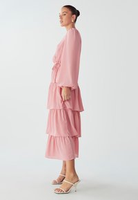 Růžové tierované šaty vyrobené z lehkého materiálu, s dlouhými rukávy, staženým pasem a vrstvenými volány, nošené s bílými sandály na podpatku.