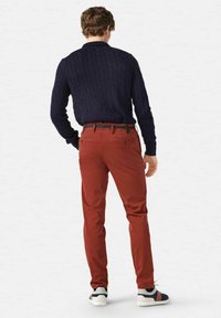 Rote Chinos mit schmaler Passform, aus Baumwolle und mit Taschen. Kombiniert mit einem navy-blauen strukturierten Pullover und mehrfarbigen Turnschuhen. Rückansicht.