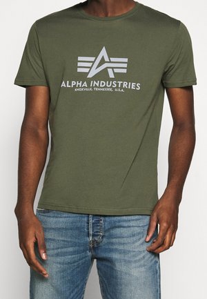 Mężczyzna w oliwkowo-zielonej koszulce Alpha Industries z logo i napisem, w połączeniu z niebieskimi jeansami, stojący na białym tle.
