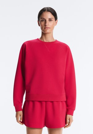 Kvinde med mørkt hår iført en matchende knaldpink sweatshirt og shorts, stående mod en lys baggrund.