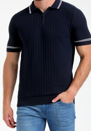 Mann trägt ein marineblau strukturiertes Kurzarm-Poloshirt mit weißen Streifenakzenten, kombiniert mit hellblauen Jeans, Hand in der Tasche.