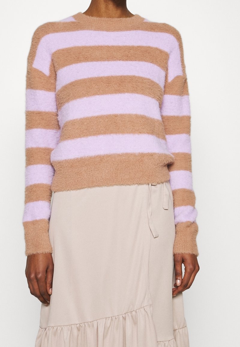 Personne portant un pull duveteux beige et violet clair rayé et une jupe portefeuille beige avec un nœud sur le côté et un ourlet à volants.