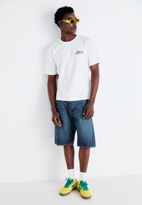 Witte katoenen t-shirt met zwart logo-print, gecombineerd met blauwe denimshorts en gele sneakers met groene accenten, gedragen met witte sokken.