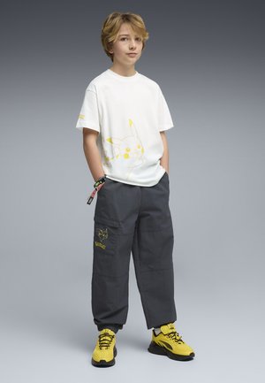 Garçon blond portant un t-shirt blanc Pikachu, un pantalon cargo noir avec un motif Pikachu, des baskets jaunes et noires, debout les mains dans les poches.