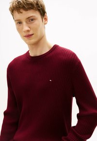 Pull en maille côtelée bordeaux avec un col rond, présentant un petit logo bleu, blanc et rouge sur la poitrine gauche. Texture lisse.