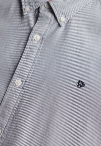 Close-up van een grijs en wit gestreepte overhemd met witte knopen en een klein geborduurd logo op de borst.