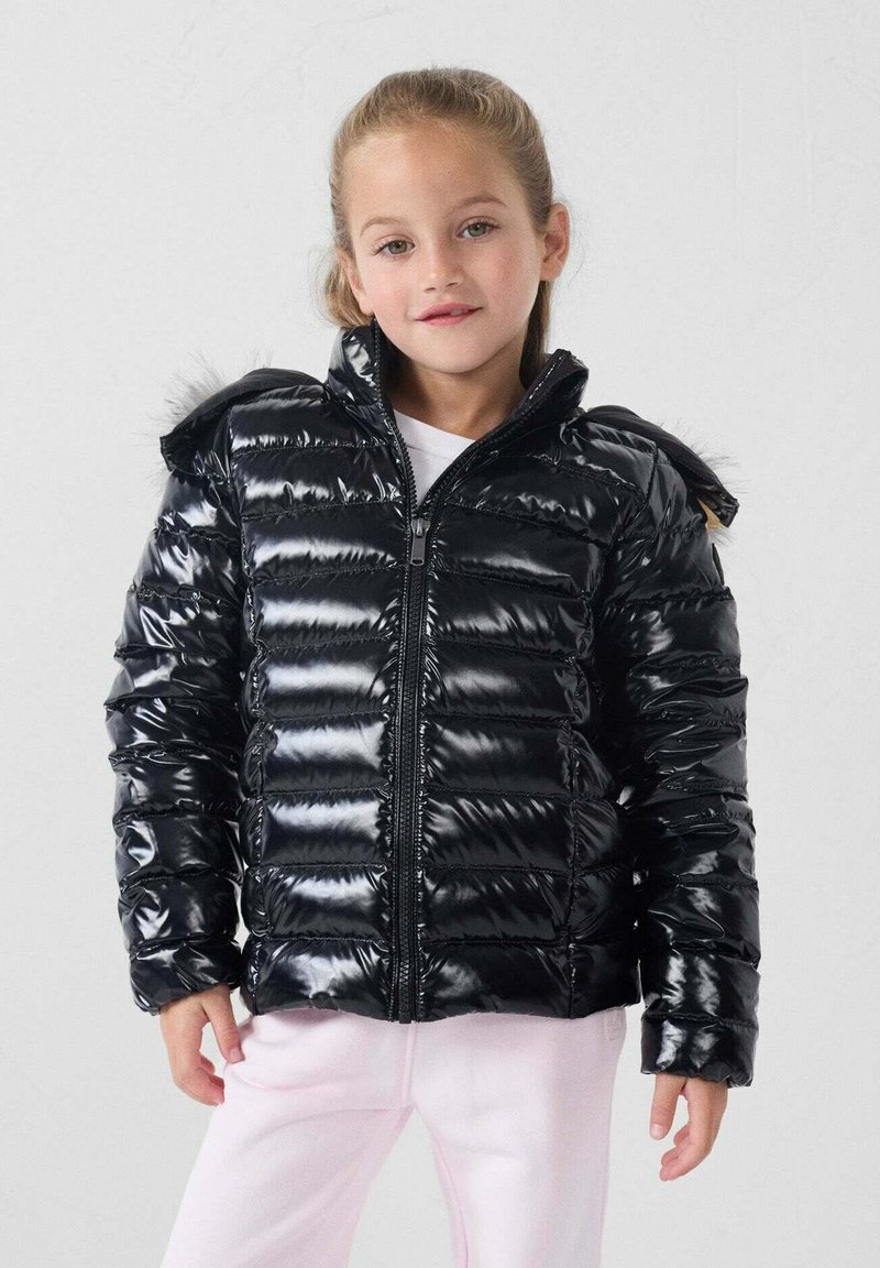 Zwarte puffjas met een glanzende, doorgestikte afwerking. Voorzien van een capuchon met bontafwerking en een rits aan de voorkant. Draagt over een wit topje en roze broek.