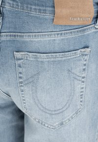 Hellblaue Jeans aus Denim mit aufgesticktem Hufeisen-Design auf der Gesäßtasche und einem hellbraunen Lederpatch mit der Aufschrift "True Religion" am Bund.