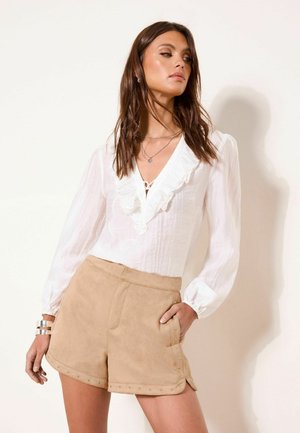 Frau trägt eine weiße, langärmelige Bluse mit Rüschen und beige Shorts mit hoher Taille und Nietenbesatz, posiert mit einer Hand in der Tasche.