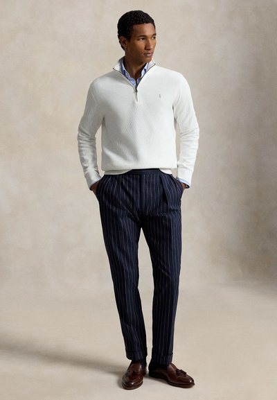 Homme portant un pull blanc à fermeture éclair quart de zip, une chemise bleue boutonnée, un pantalon à fines rayures bleu marine et des mocassins marron à glands, debout, les mains dans les poches.