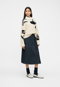 Γυναίκα που φοράει εκρού πουλόβερ με μαύρα αφηρημένα μοτίβα, λευκό πουκάμισο με γιακά, σκουρόχρωμη μίντι φούστα από τζιν, σκουρόχρωμες κάλτσες και λευκά loafers.