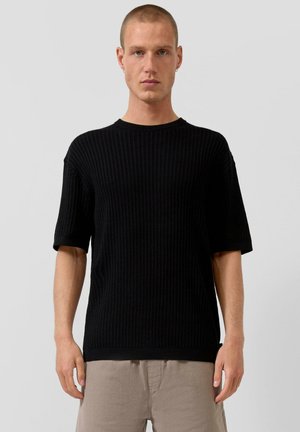 Junger Mann mit kurzen Haaren, der silberne Creolen, einen schwarzen kurzärmeligen, gerippten Strickpullover und hellbeige Hosen trägt, vor einem grauen Hintergrund stehend.