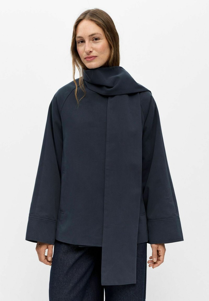 Femme portant une veste bleu marine foncé ample avec de larges manches et une longue écharpe intégrée qui pend devant.
