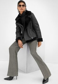 Frau in schwarzer Lammfelljacke, gemusterten Schlaghosen, gepunkteten High Heels und Sonnenbrille, posiert sitzend auf einem weißen Würfel.