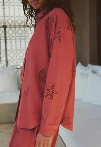 Punane linane särk, millel on varrukatel ja äärisel lillemustriga lõiked. Omab klassikalist kraega ja nööbitavat sulgemist. Sile tekstuur.