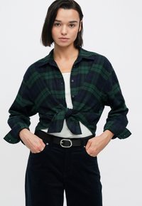 Πουκάμισο από φανέλα με πράσινο και ναυτικό καρό δεμένο στη μέση, με γιακά και κλείσιμο με κουμπιά, σε συνδυασμό με σκούρο παντελόνι και μαύρη ζώνη.