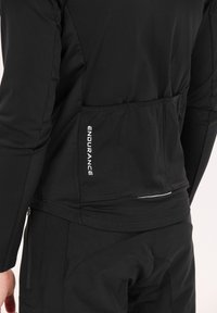 Veste de sport noire en tissu texturé, avec une poche latérale et des détails réfléchissants. Logo imprimé verticalement sur le côté gauche.