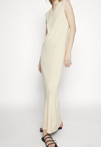 Robe maxi sans manches en tricot beige clair avec un décolleté en V et des détails côtelés. Présente un ourlet évasé et est portée avec des sandales noires à lanières.