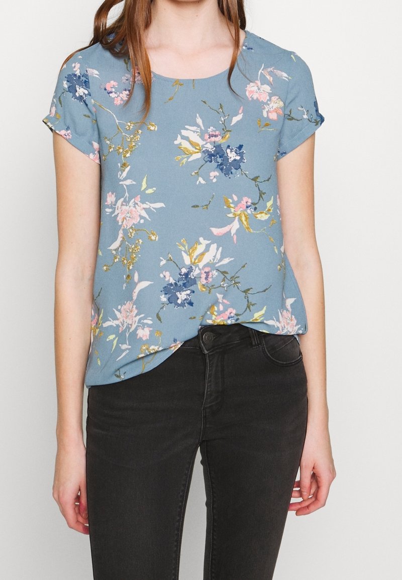 Blusa floral en azul claro, con mangas cortas, un escote redondeado y patrones florales multicolores con acentos dorados.