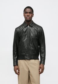 Μαύρο δερμάτινο μπουφάν με ανάγλυφα σχέδια, όρθιο γιακά, μπροστινό φερμουάρ και ριμπ τελείωμα. Φορεμένο πάνω από ανοιχτόχρωμο παντελόνι.