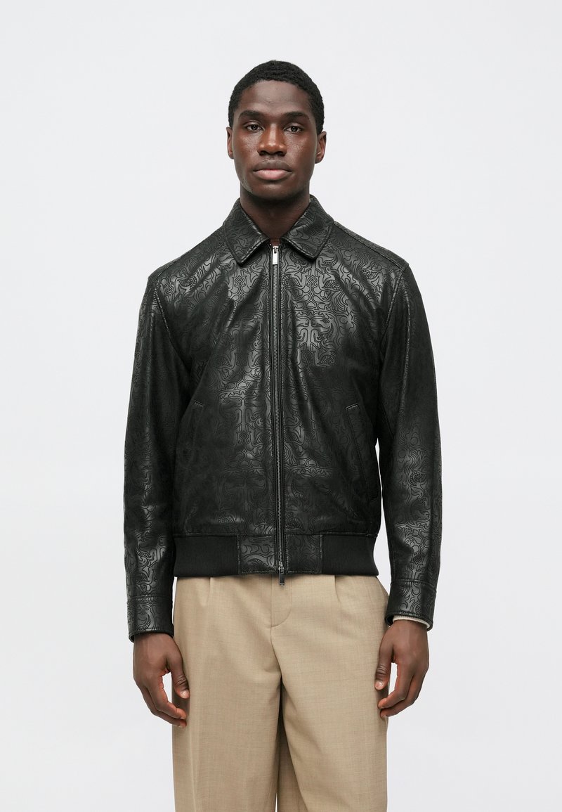 Μαύρο δερμάτινο μπουφάν με ανάγλυφα σχέδια, όρθιο γιακά, μπροστινό φερμουάρ και ριμπ τελείωμα. Φορεμένο πάνω από ανοιχτόχρωμο παντελόνι.