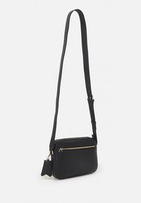 Černá křížová taška vyrobená z texturované kůže. Obsahuje zipovou přední kapsu, nastavitelný popruh a dekorativní štítek s ostrými akcenty.
