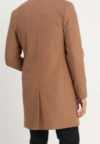 Cappotto in misto lana marrone, di lunghezza media, con una forma dritta e cuciture minime. Presenta maniche lunghe e una texture morbida.