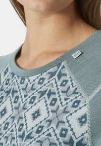 Licht teal lange mouw top met een geometrisch patroon in donkerder teal. Heeft een geribbelde halslijn en een stoffen label op de schouder.