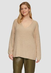 Beige gebreide trui met V-hals, oversized pasvorm, lange mouwen en een gestructureerde afwerking. Gedragen over donkergroene leren broeken.