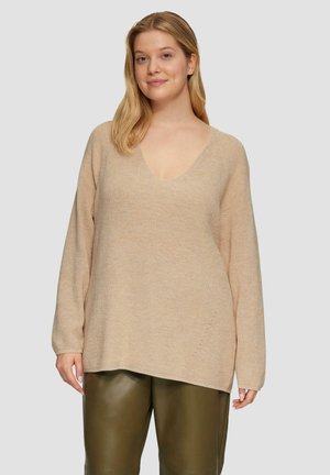 Beige gebreide trui met V-hals, oversized pasvorm, lange mouwen en een gestructureerde afwerking. Gedragen over donkergroene leren broeken.