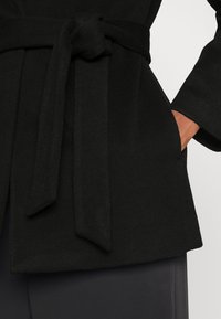 Abrigo negro con cinturón, confeccionado en una tela suave y texturizada. Presenta un lazo ancho y bolsillos, un diseño entallado y una silueta relajada.