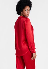 Chemisier en satin rouge avec un col, des manches longues et des poignets boutonnés, présentant une texture lisse et un ourlet légèrement courbé.