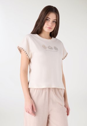 Giovane donna indossa una maglietta beige a maniche corte con maniche a frange e una stampa con la scritta "Prenditi un momento per respirare", abbinata a pantaloni beige chiaro.