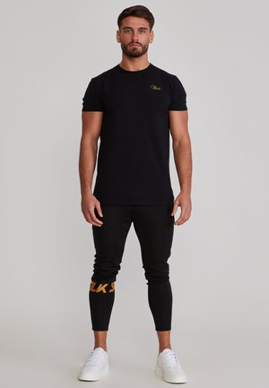 Mężczyzna stojący przodem, ubrany w czarną koszulkę z krótkim rękawem, czarne spodnie z pomarańczowym napisem na jednej nodze oraz białe trampki.