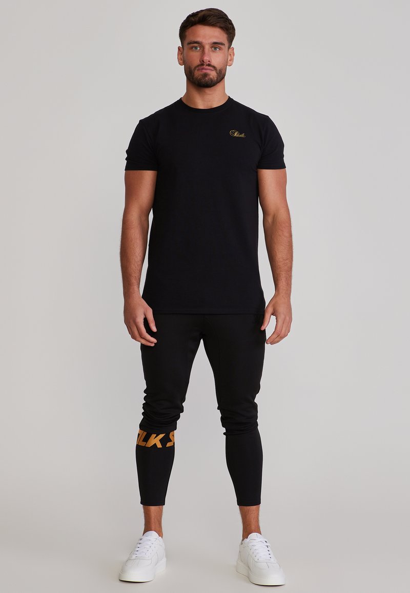 Muž stojaci čelom dopredu, oblečený v čiernom tričku s krátkym rukávom, čiernych nohaviciach s oranžovým textom na jednom kurze a bielych teniskách.