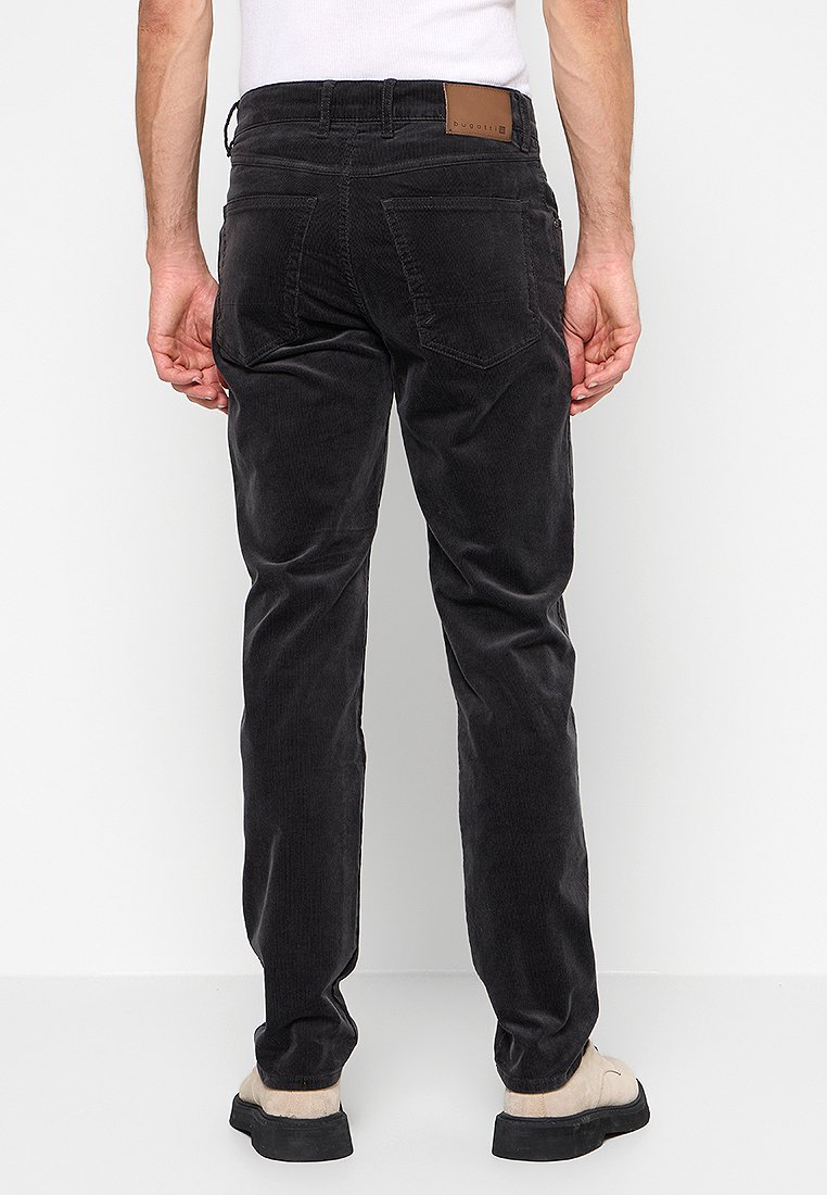 Mann in schwarzen Cordhosen, mit einem in die Hose gesteckten weißen Hemd und beigen Schuhen, steht mit dem Rücken zu einer schlichten weißen Wand.